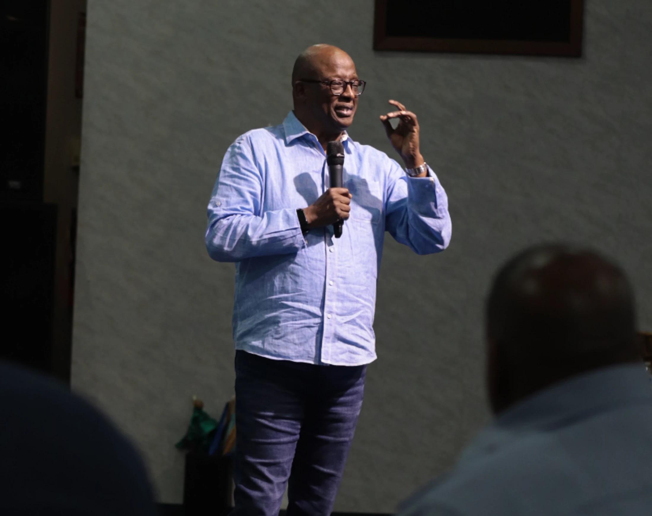 A man in a light blue shirt and glasses stands holding a microphone, speaking and gesturing to an audience whose backs are visible in the foreground.