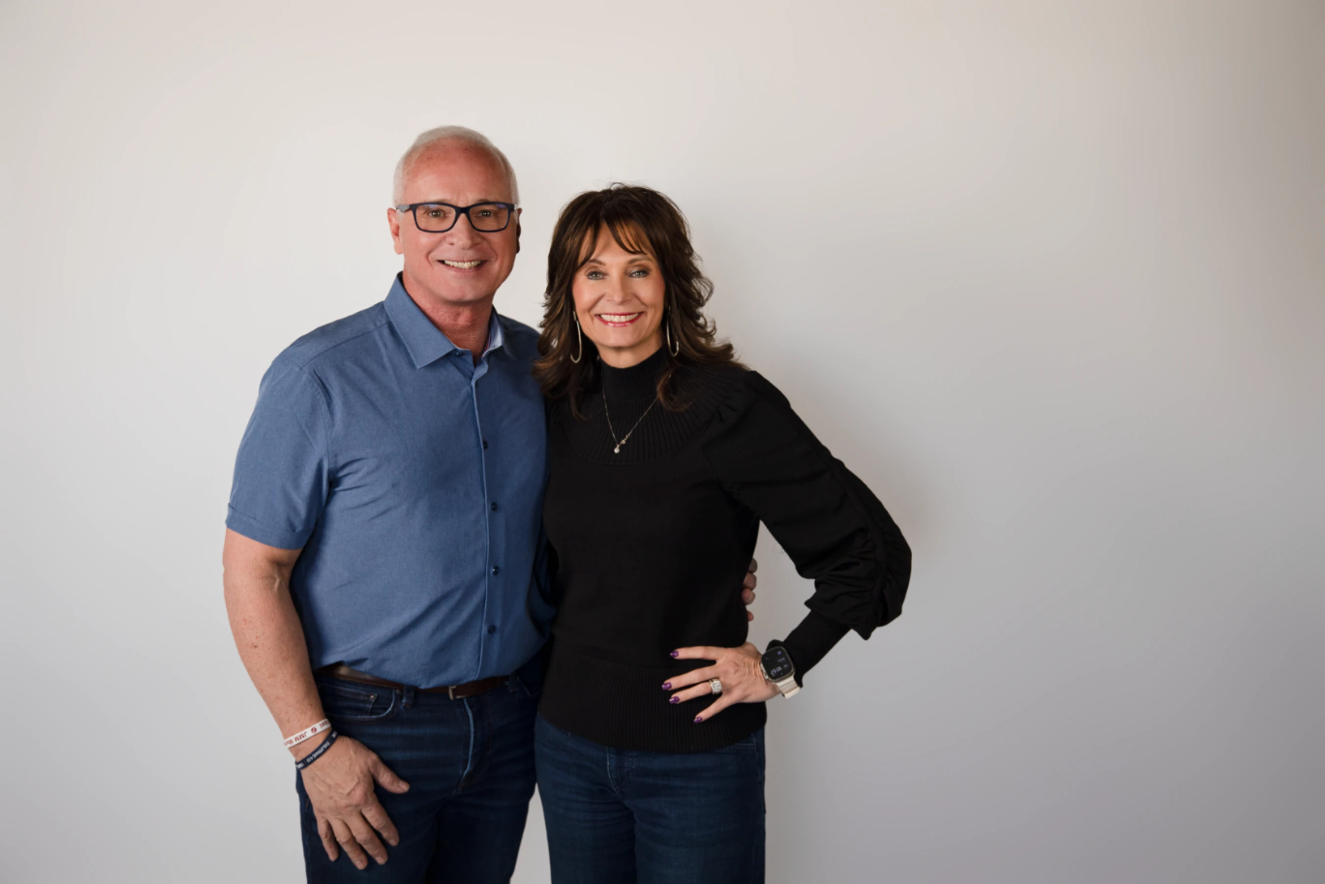 A smiling man and woman pose together against a plain white background. The man wears glasses and a blue shirt, and the woman wears a black sweater.