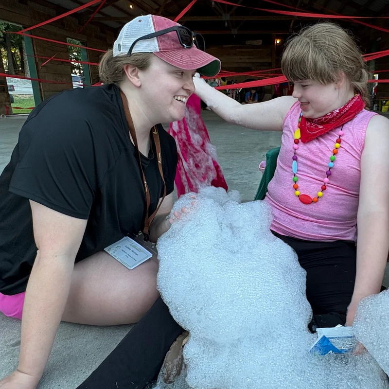 A female camp counselor and a young girl smile at each other while sitting on the ground playing with large piles of white foam bubbles under a pavilion.