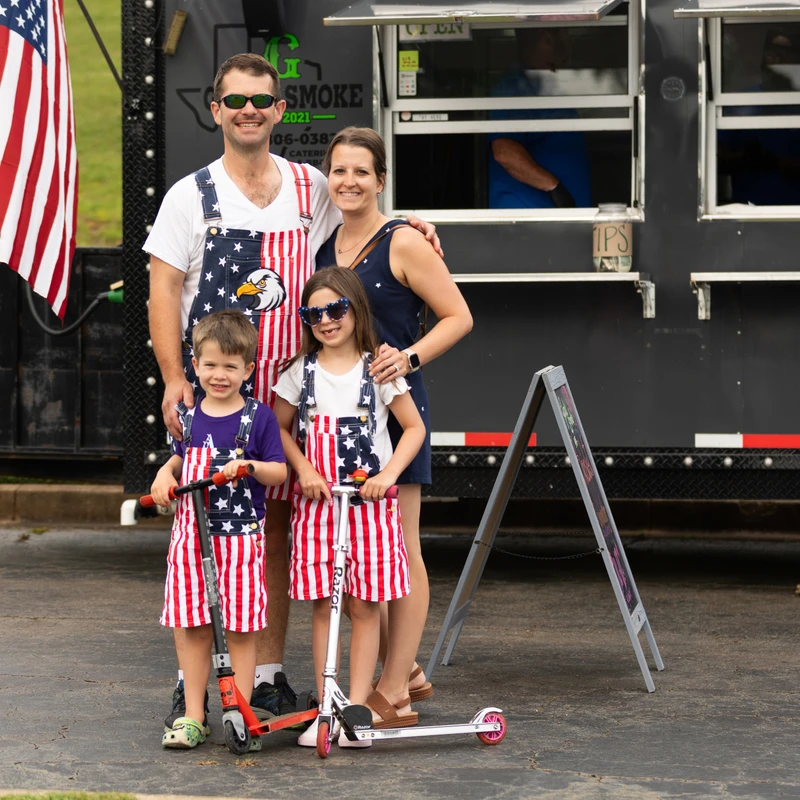 A smiling family of four wearing patriotic stars-and-stripes overalls poses in front of a black food truck, with the two children holding scooters.