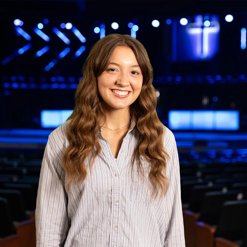 A young woman with long brown hair smiles while standing in an empty auditorium. The stage behind her is lit with blue lights and shows a cross.