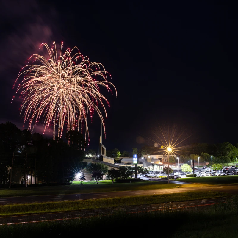 A long exposure night photograph of a golden and red firework bursting in the dark sky above an illuminated road and a building with a full parking lot.