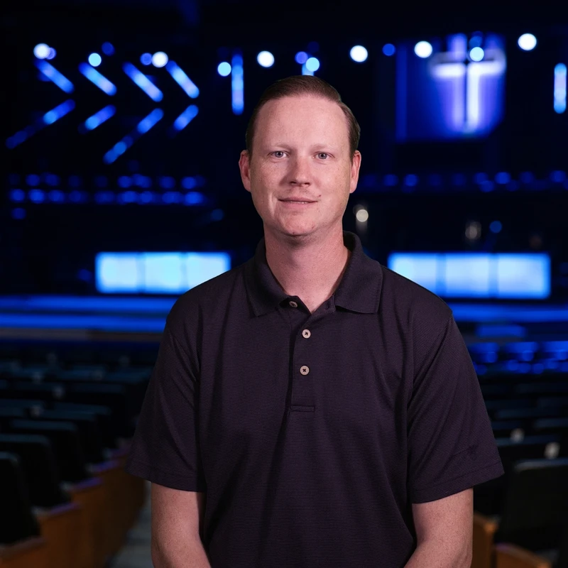 A headshot of a man in a dark polo shirt standing in a large, dark auditorium. The stage behind him is lit with blue lights and features a glowing cross.