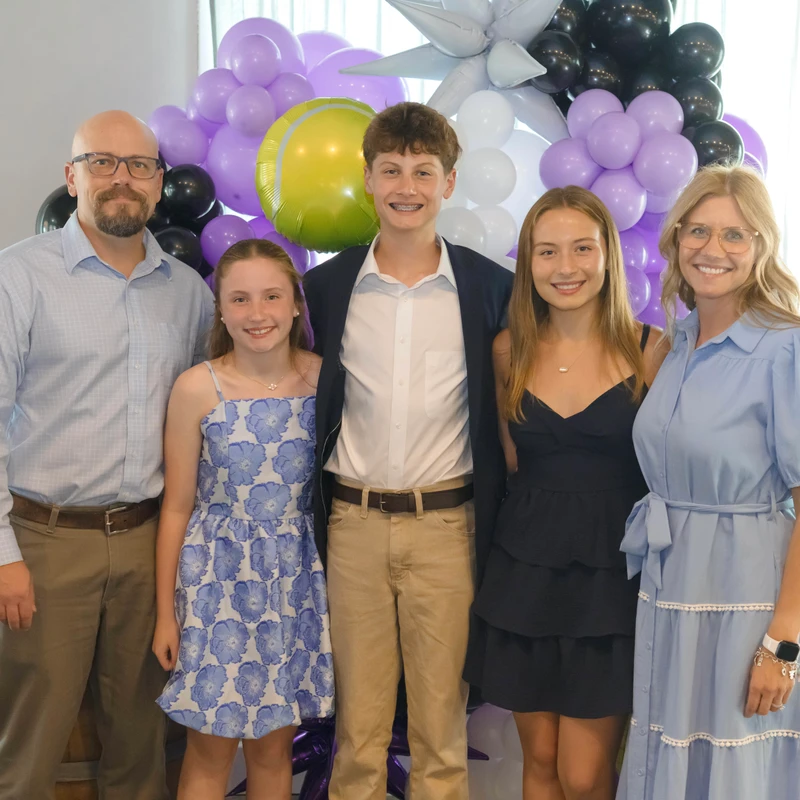 A smiling family of five, with parents and three children, stands together for a photo in front of a backdrop of purple, black, and tennis ball balloons.