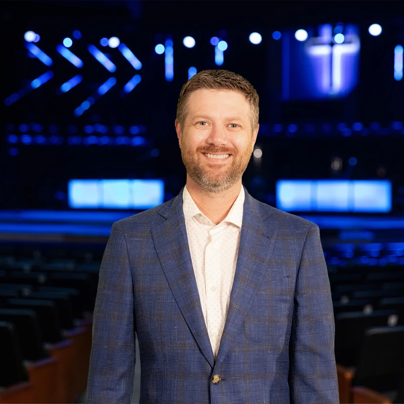 A man with a beard, wearing a blue blazer, smiles while standing in an empty auditorium. In the background, a stage is lit with blue lights and a cross.