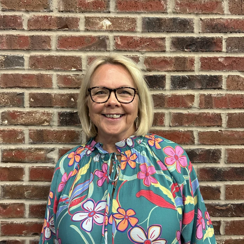 A headshot of a smiling blonde woman with glasses wearing a colorful floral blouse, standing in front of a red brick wall.
