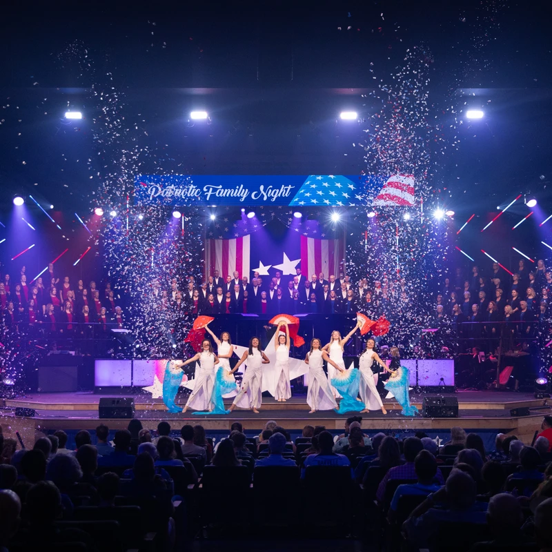 Dancers and a choir perform on a brightly lit stage during a patriotic show as red, white, and blue confetti falls, viewed by a large audience.