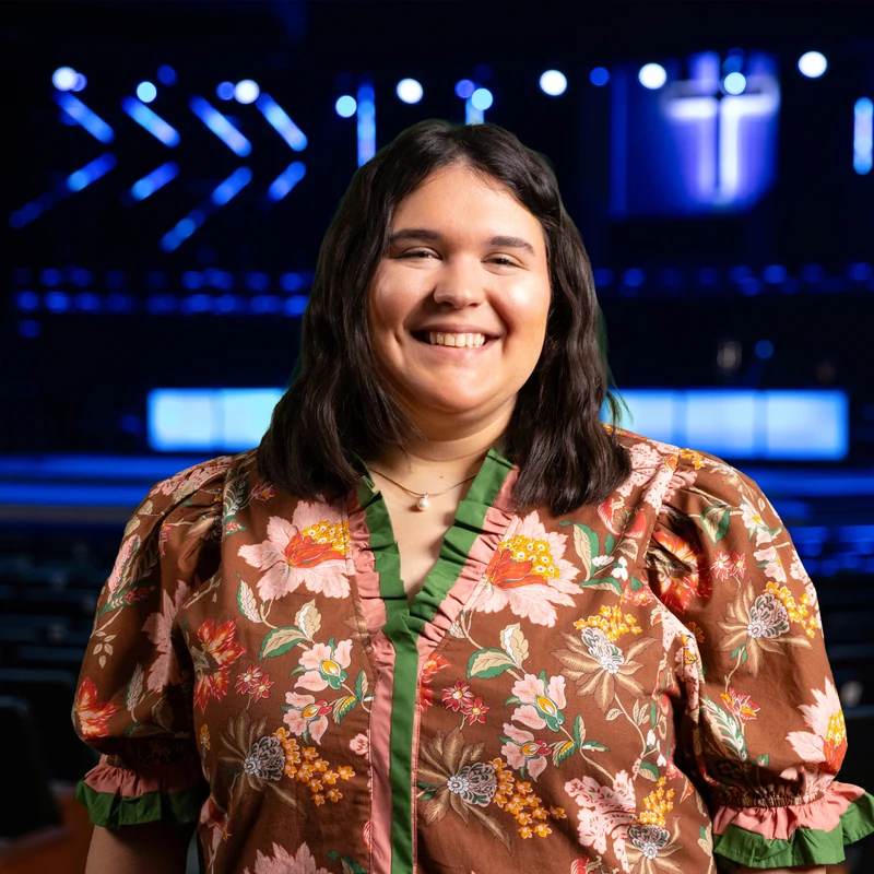 A young woman with dark hair smiles warmly. She wears a floral blouse and stands in an auditorium with a blue-lit stage and a glowing cross behind her.