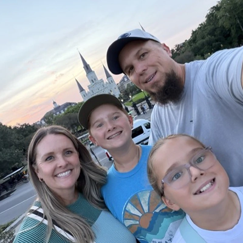 A smiling family of four takes a selfie outdoors at sunset. In the background, the spires of St. Louis Cathedral rise against a pink and blue sky.