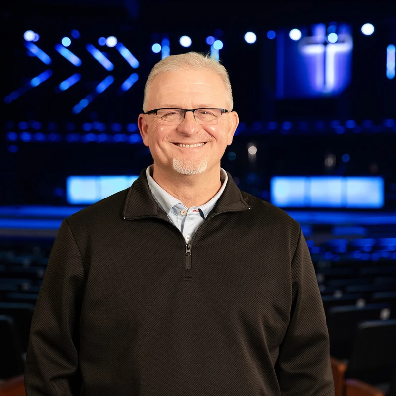 A smiling, middle-aged man with gray hair, a goatee, and glasses stands in an auditorium. Behind him, a stage is lit with blue lights and a visible cross.