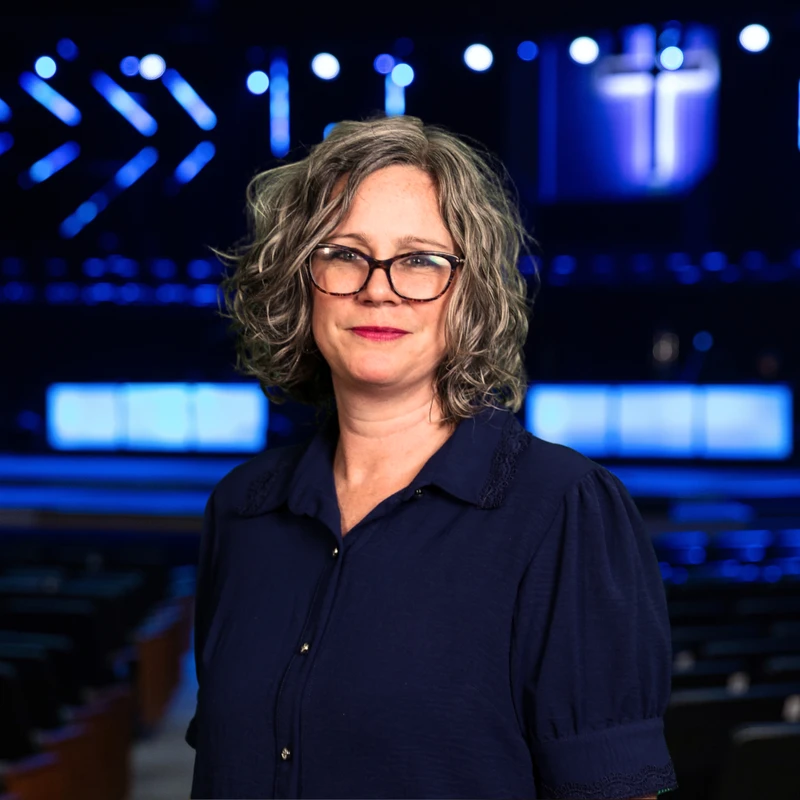 A professional headshot of a woman with curly gray hair and glasses, smiling in front of a stage with vibrant blue lights and a glowing cross in the background.