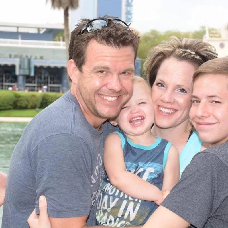 A smiling family with two parents, a teenage son, and a laughing toddler embrace for a photo outdoors with a body of water in the background.