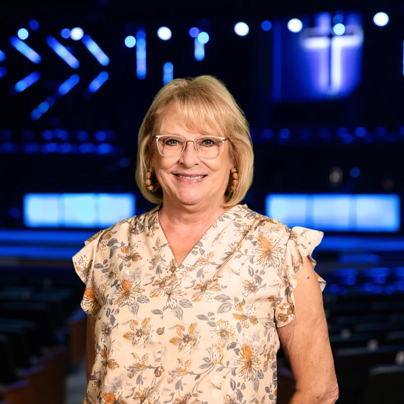 A smiling woman with blonde hair and glasses, wearing a floral blouse, stands in front of a dark stage with blurred blue lights and a glowing cross.