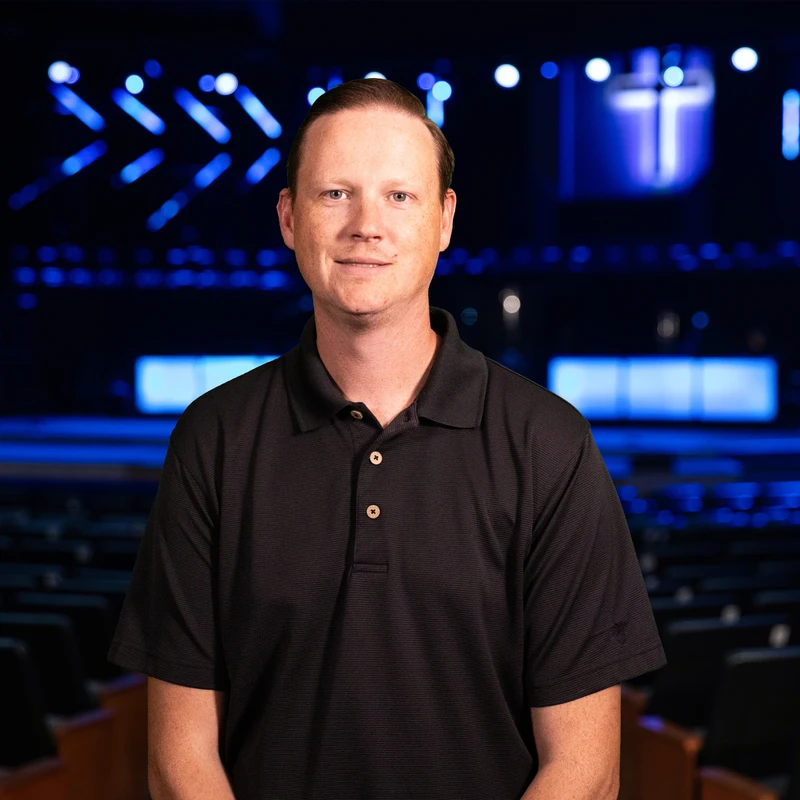 A man in a black polo shirt stands in front of an empty auditorium with a stage lit by blue lights and a cross in the background.
