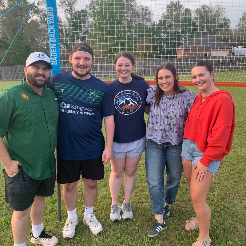 Five friends, two men and three women, stand smiling in front of a backyard volleyball net on a grassy lawn, dressed in casual clothing.