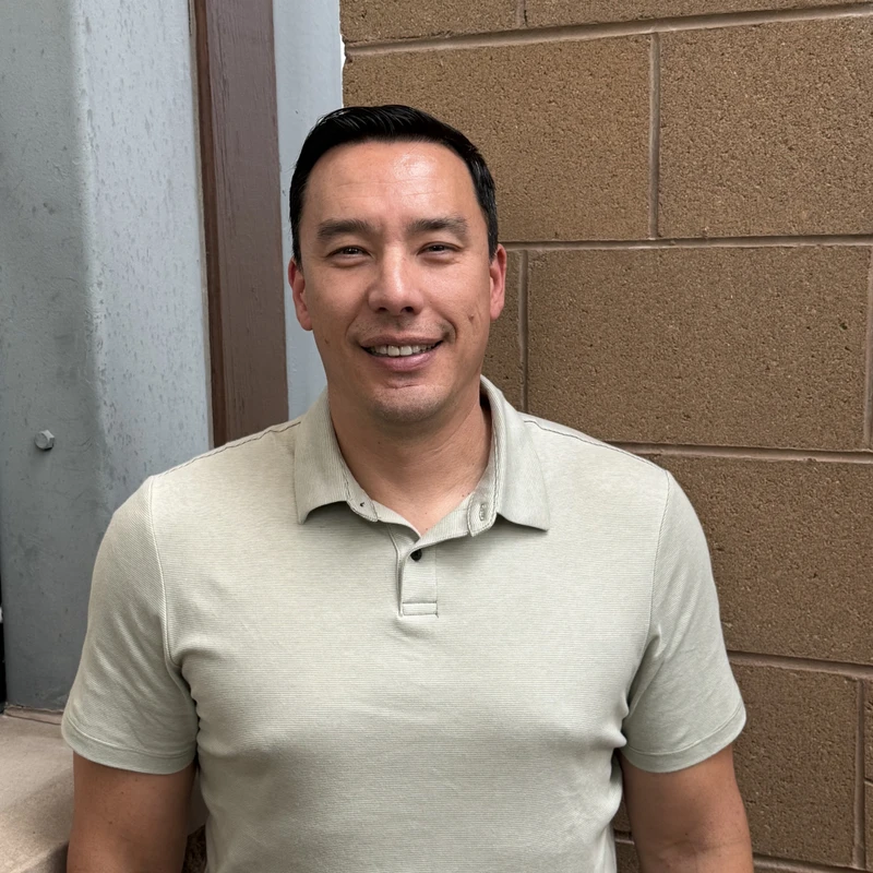 A man with short dark hair in a light-colored polo shirt smiles for the camera while standing in front of a brown brick wall.