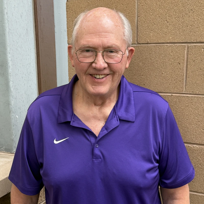 A friendly older man with glasses and white hair, wearing a purple polo shirt, smiles for a portrait against a tan brick wall.