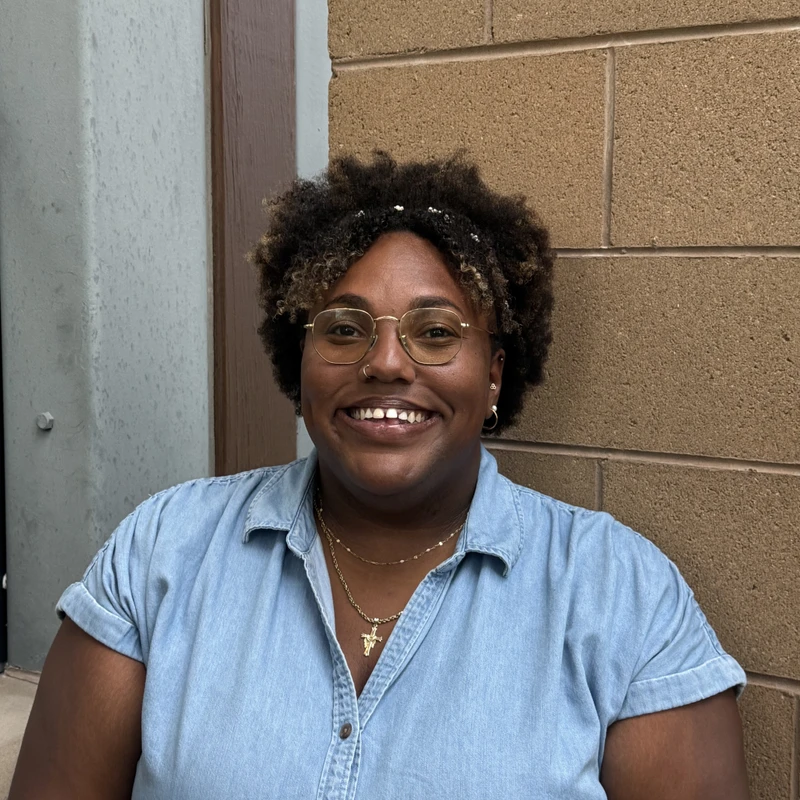 A head and shoulders portrait of a smiling Black woman with curly hair and glasses, wearing a denim shirt and standing in front of a brick wall.