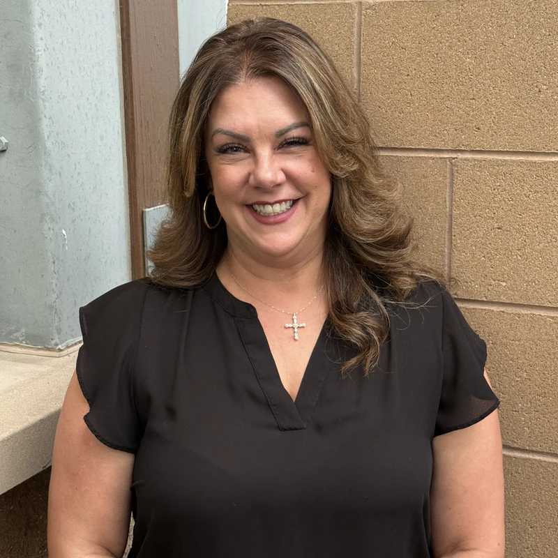 A woman with styled, highlighted brown hair smiles at the camera. She wears a black top, a cross necklace, and hoop earrings, posing by a brick wall.