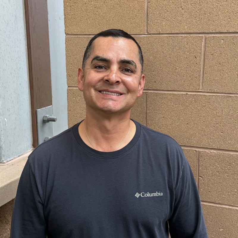 A medium shot of a smiling man with short dark hair, wearing a dark gray long-sleeved Columbia shirt. He is standing in front of a tan brick wall.