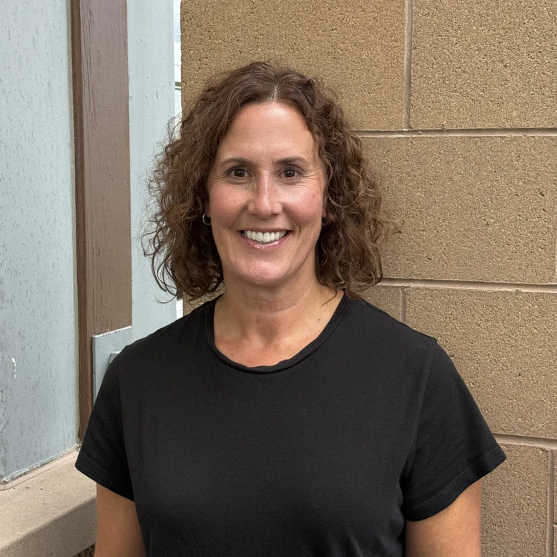 A friendly-looking woman with curly, shoulder-length brown hair smiles for the camera. She is wearing a black t-shirt and stands against a brick wall.