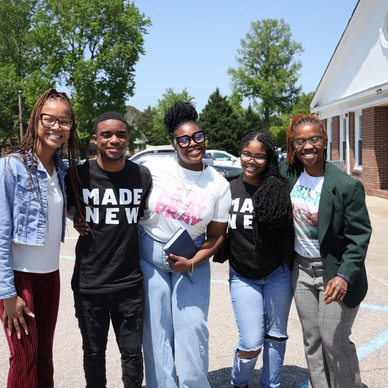 A group of five young Black people stand arm-in-arm, smiling for the camera in a parking lot on a bright, sunny day with trees in the background.