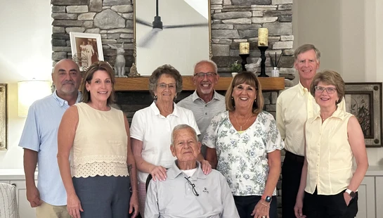A group of eight family members smile for a portrait in a living room. An elderly man is seated in front of a stone fireplace with others standing around him.