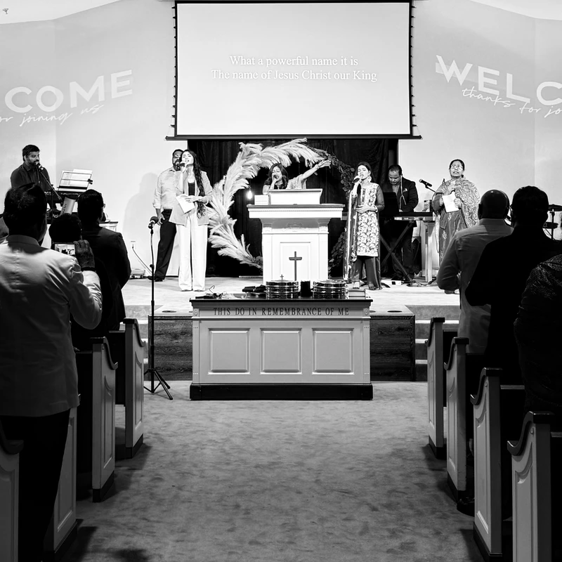 A black and white photograph of a church service, showing the congregation facing a stage where a band is performing and lyrics are displayed on a screen.