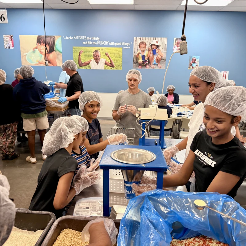 A diverse group of smiling volunteers of all ages wear hairnets while packing food into bags at an assembly line for a hunger relief organization.