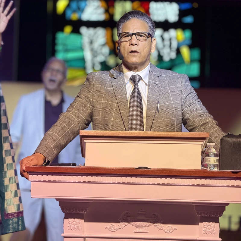 A man in a grey plaid suit and glasses stands at a pink podium, speaking with an intense expression. Another man sings in the background on a stage.