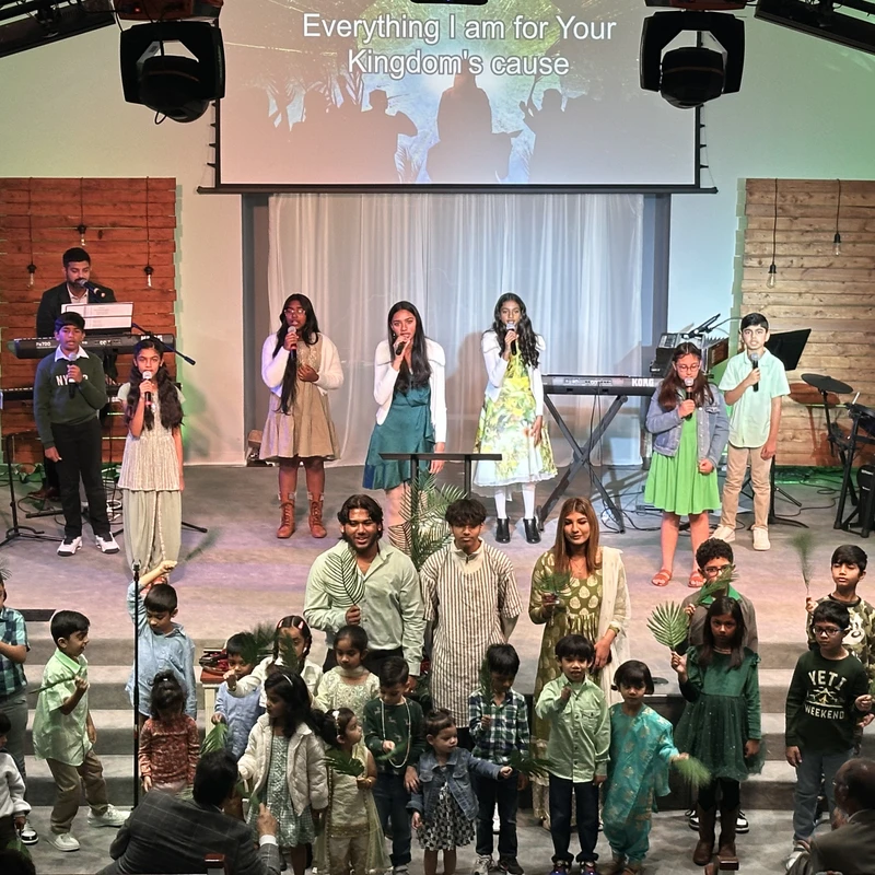 A large group of children and adults performing on a church stage, singing and holding palm fronds, with a band and lyrics projected on a screen behind them.