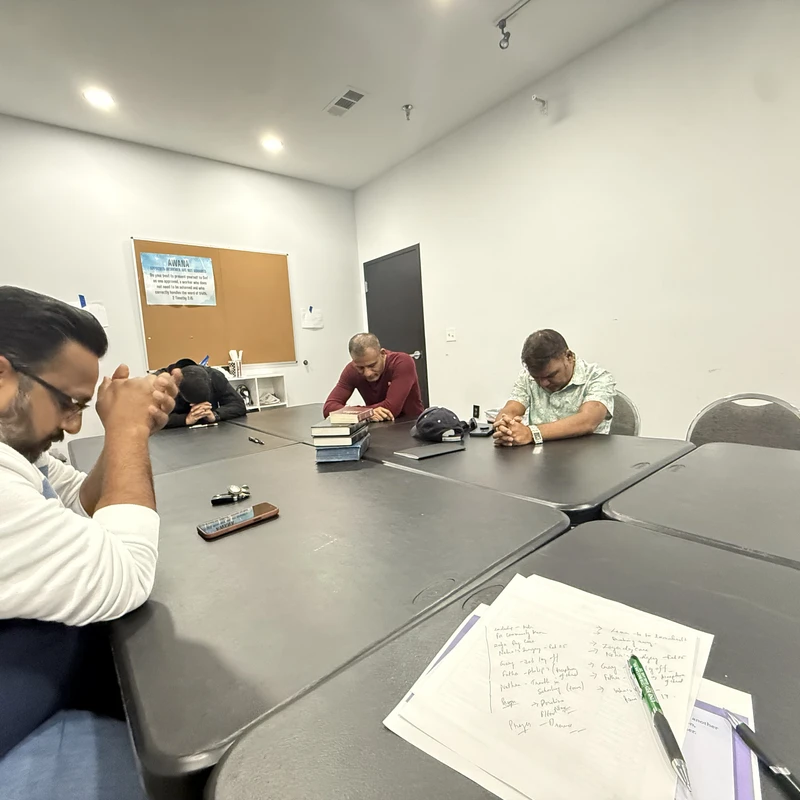 Several men sit around a large black table in a classroom, their heads bowed and hands clasped in a moment of prayer or reflection.