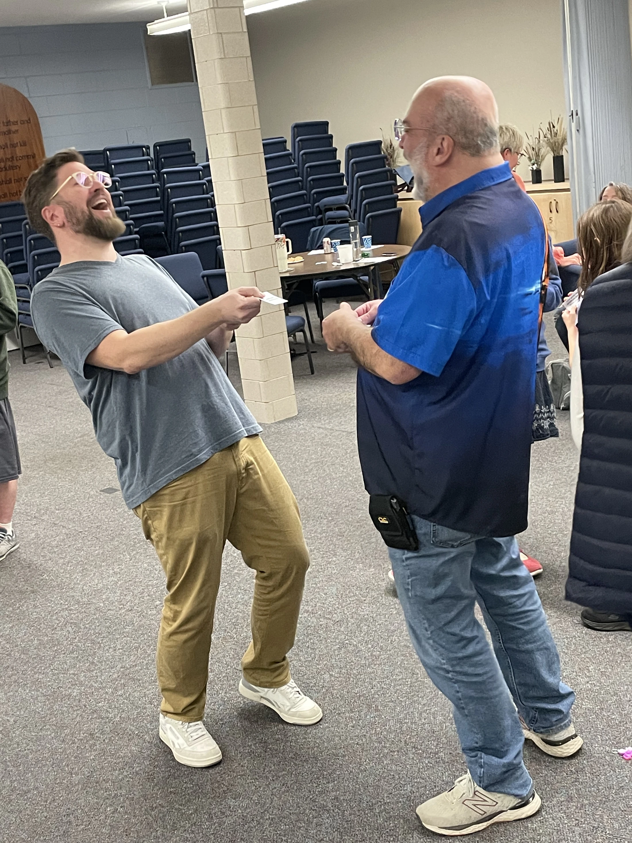 A younger man with a beard and glasses laughs as he playfully pulls a small card from an older man's hand in a large room with stacked chairs.