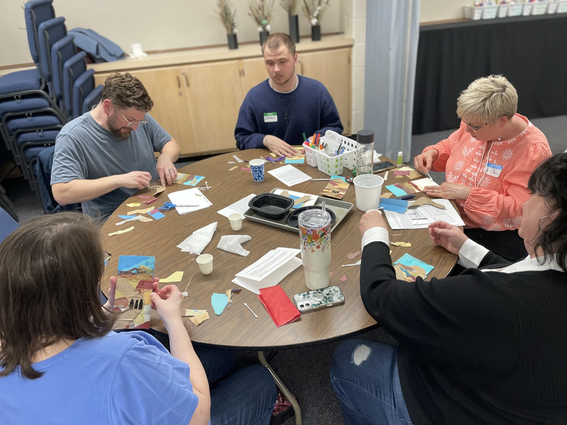 Five adults sit around a circular wooden table, concentrating on creating collages with various pieces of colored paper, scissors, and glue.