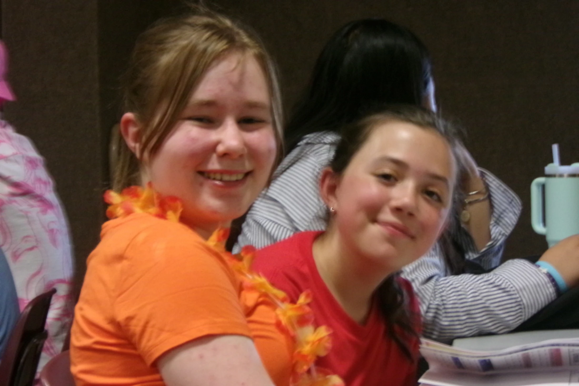 Two young girls sit next to each other and smile for the camera. The girl on the left wears an orange shirt and a flower lei around her neck.