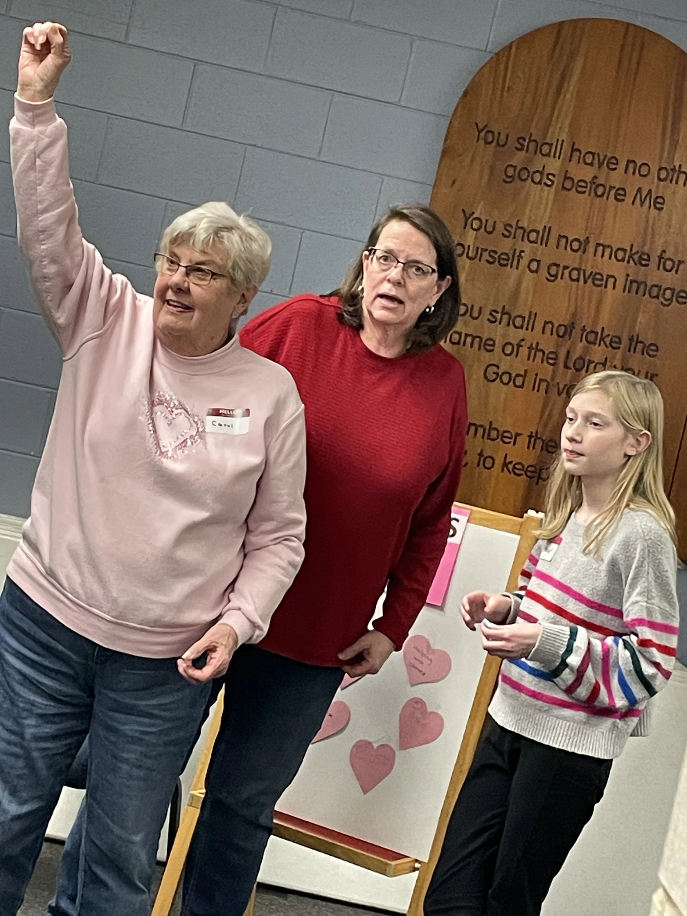 An older woman in a pink sweater raises her fist enthusiastically, standing next to another woman and a young girl in front of a Ten Commandments plaque.