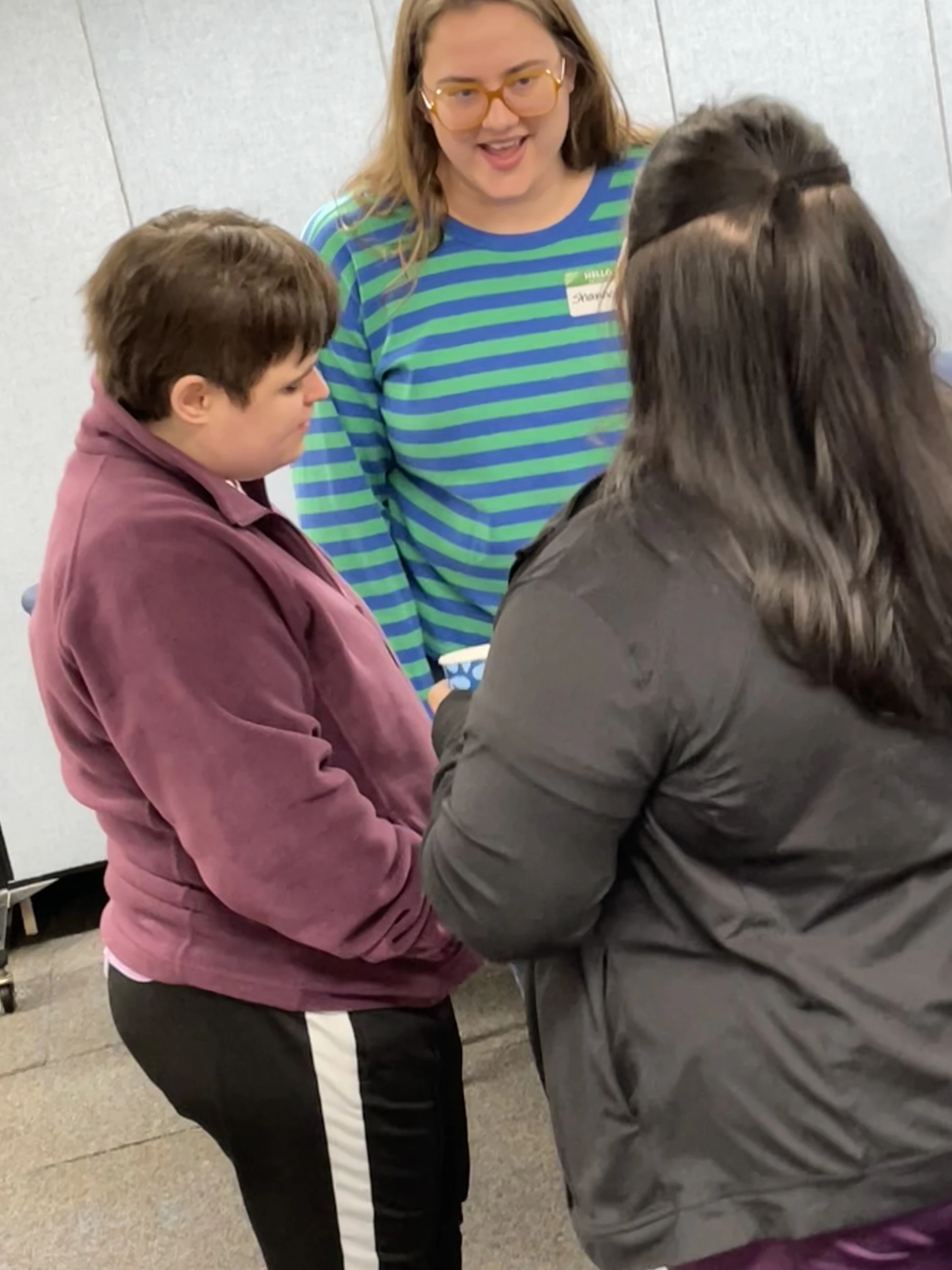 Three women stand together talking. The woman in the center smiles, wearing glasses and a blue and green striped shirt, looking at the other two.