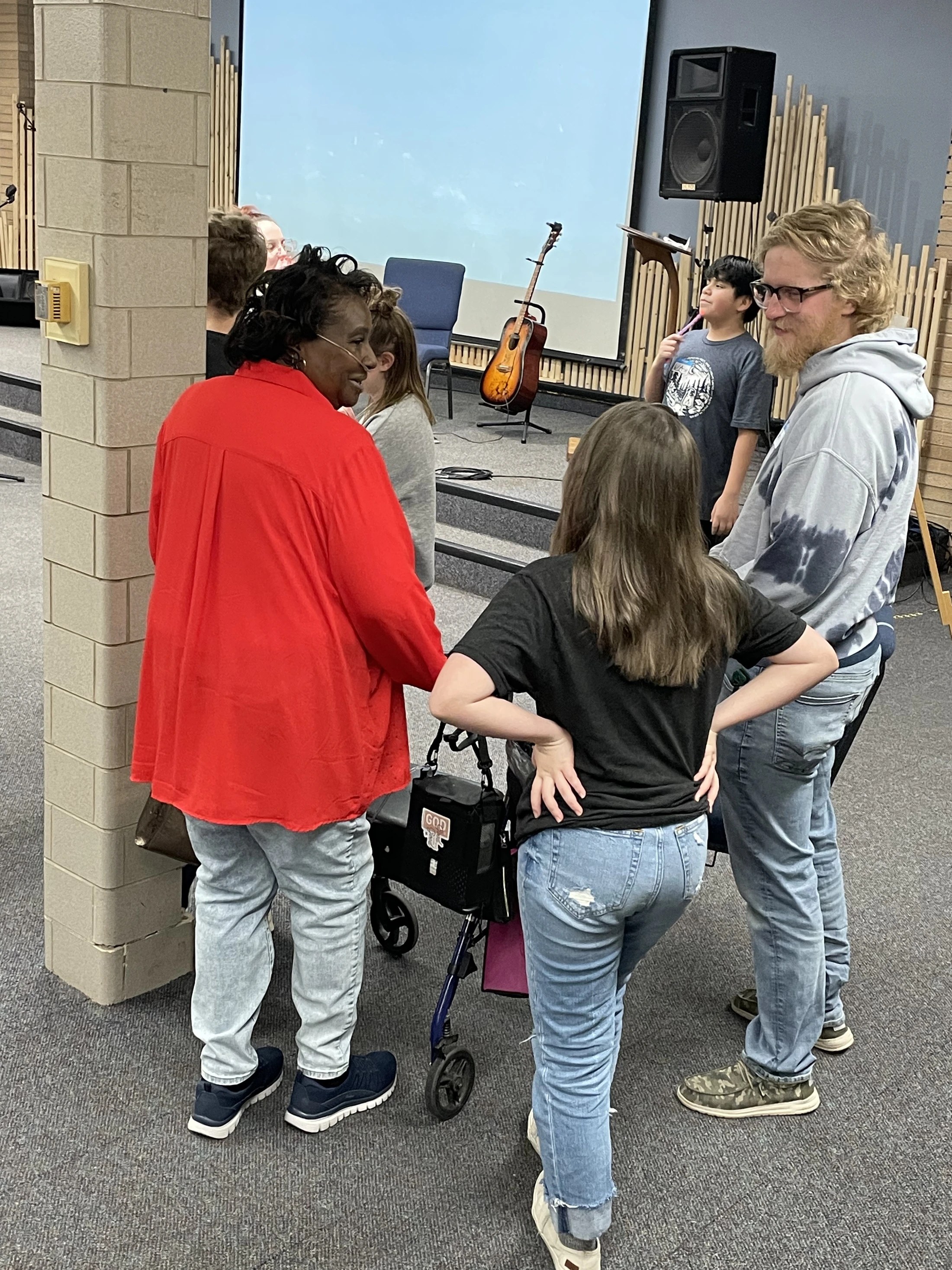 A diverse group of people chat in a circle in what appears to be a church hall. A woman in red with a walker talks with a young man and a girl.