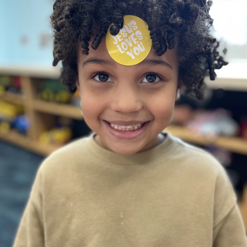 A close-up portrait of a happy young child with curly hair, smiling at the camera with a yellow 'Jesus Loves You' sticker on their forehead.