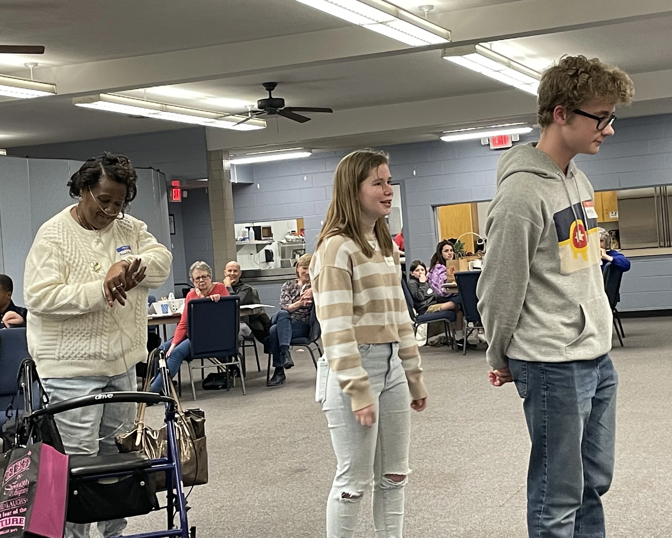 An older woman with a walker, a teenage girl, and a teenage boy stand in front of a seated audience in a community room during an event.