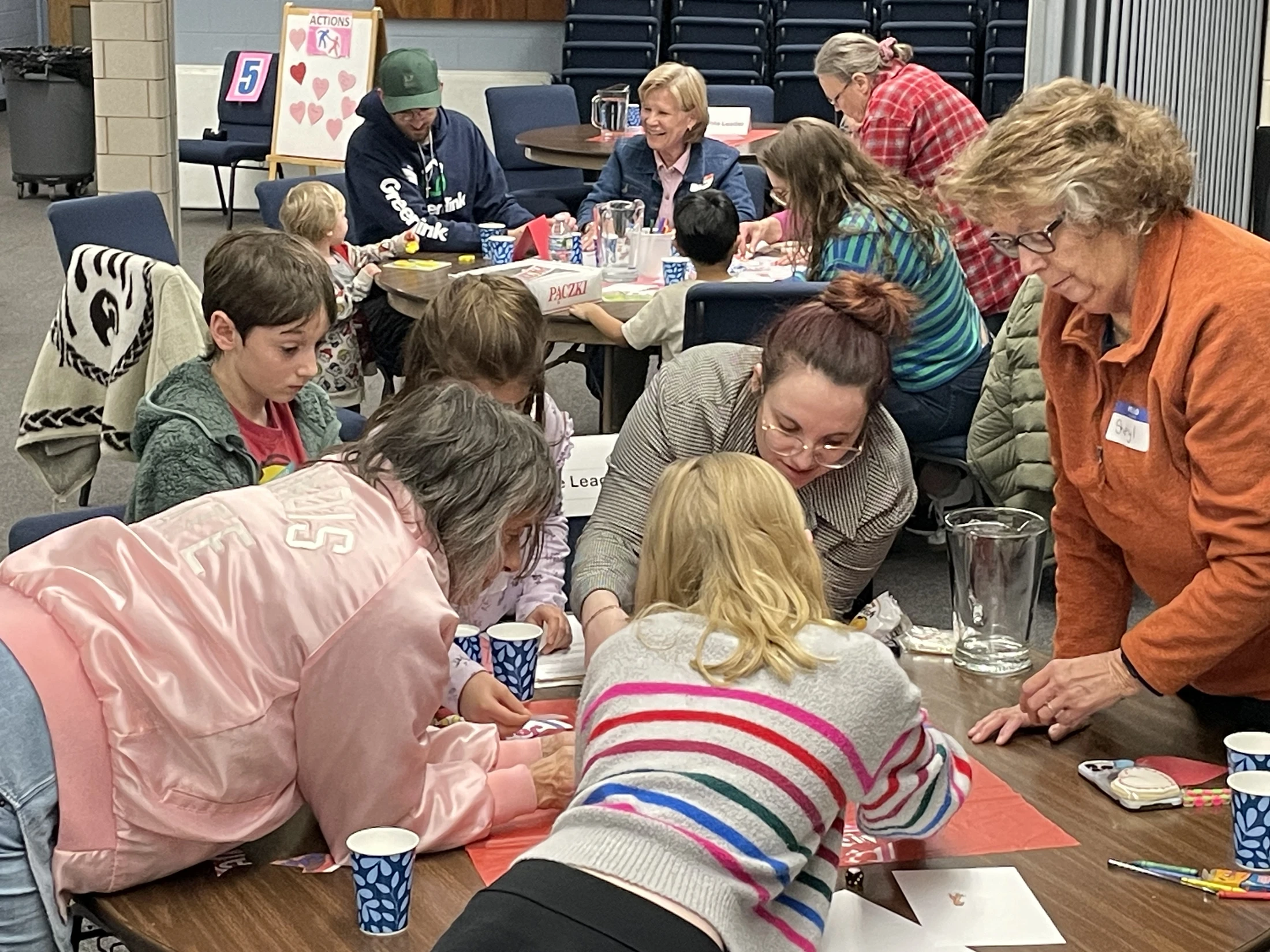A large, diverse group of adults and children gathered around tables indoors, working together on an arts and crafts project in a community setting.
