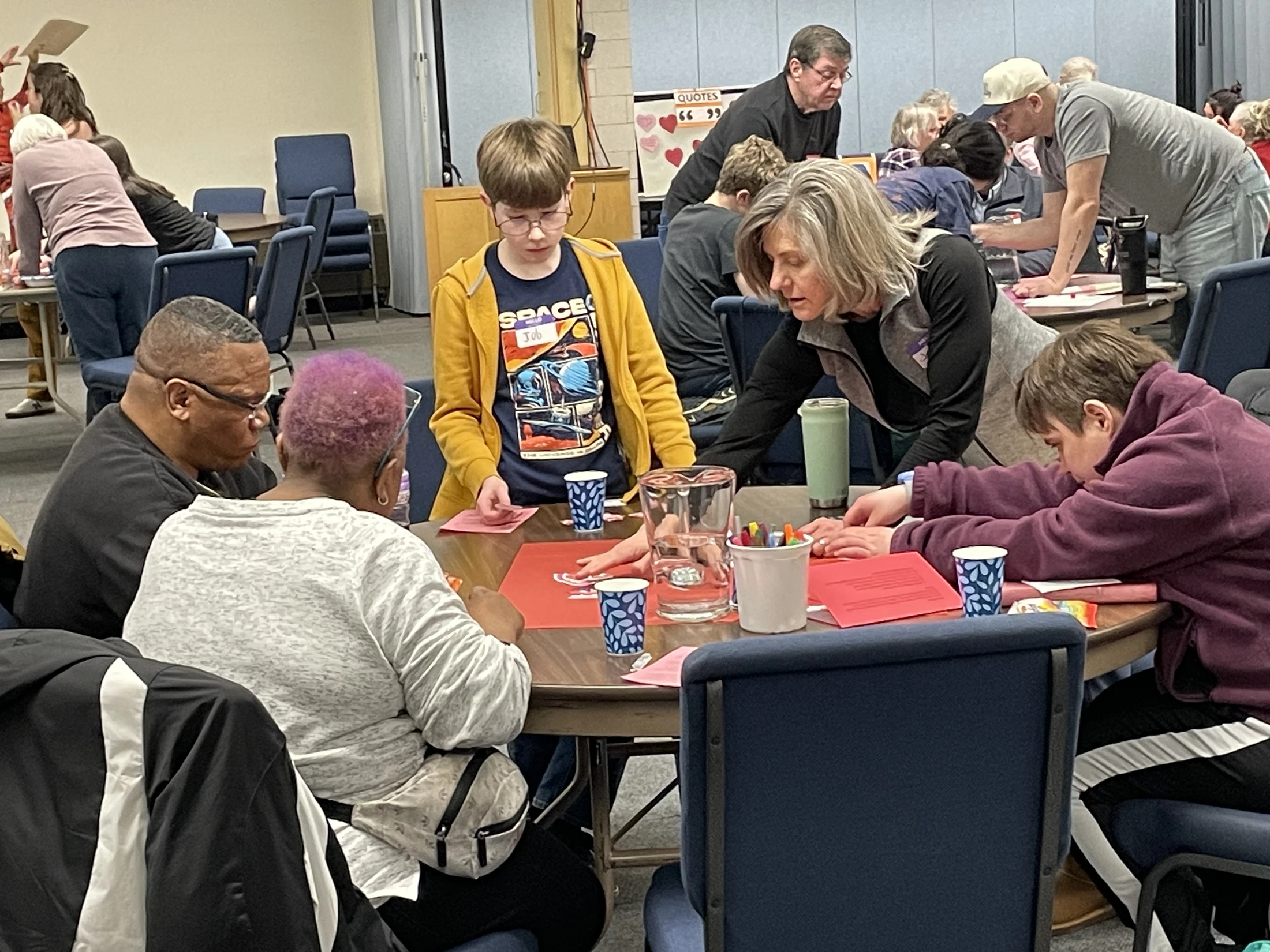 A diverse group of people of different ages sitting around a table, working together on a craft project in a large room full of other participants.