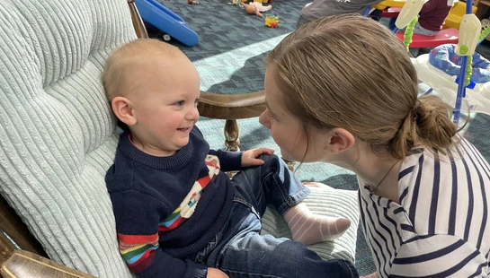 A smiling baby boy sits in a cushioned rocking chair while an older girl leans in close, looking at him face-to-face in a colorful children's playroom.