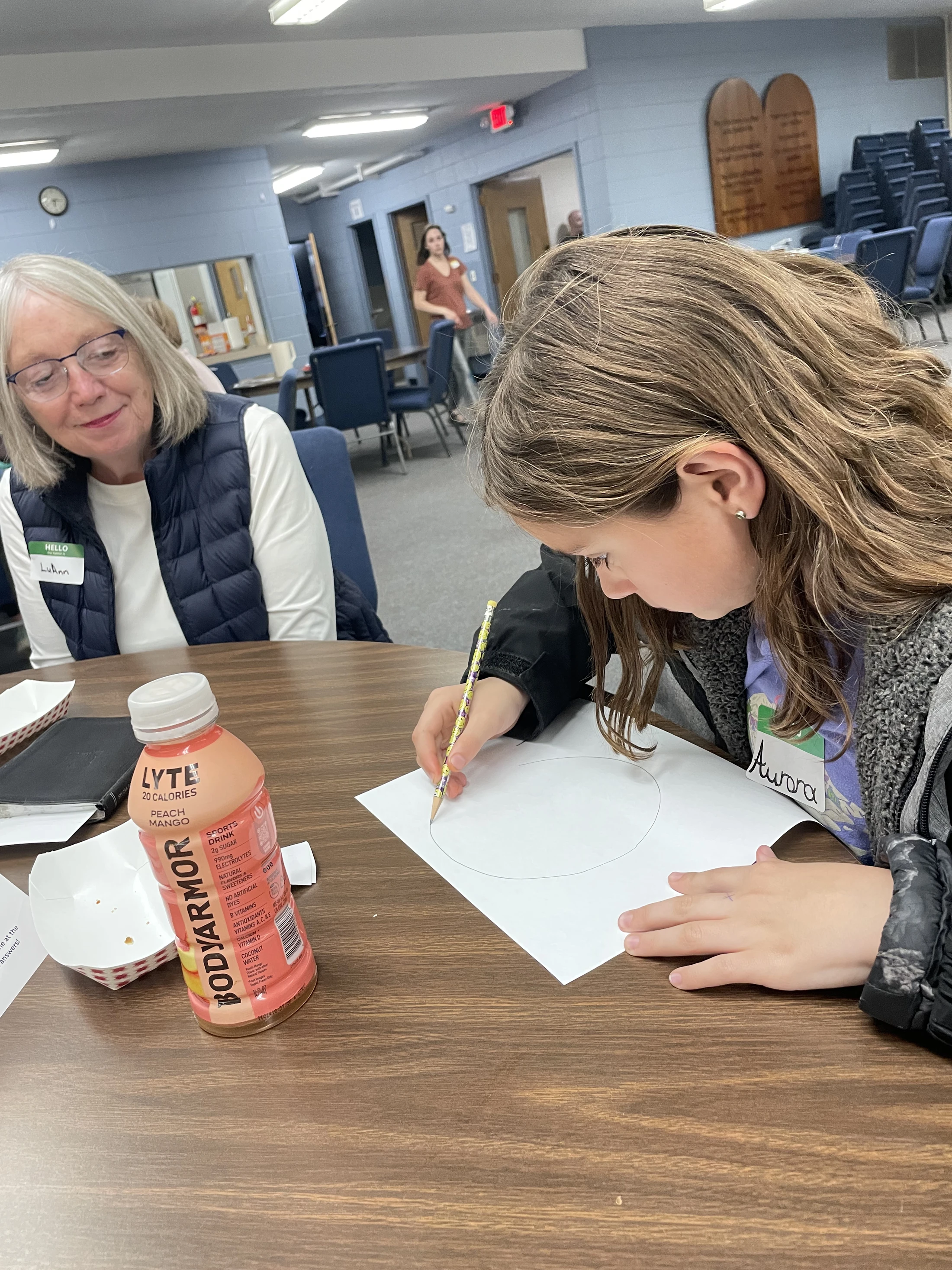 A young girl with long brown hair draws a circle on paper with a pencil while an older woman with glasses watches her with a warm smile at a wooden table.