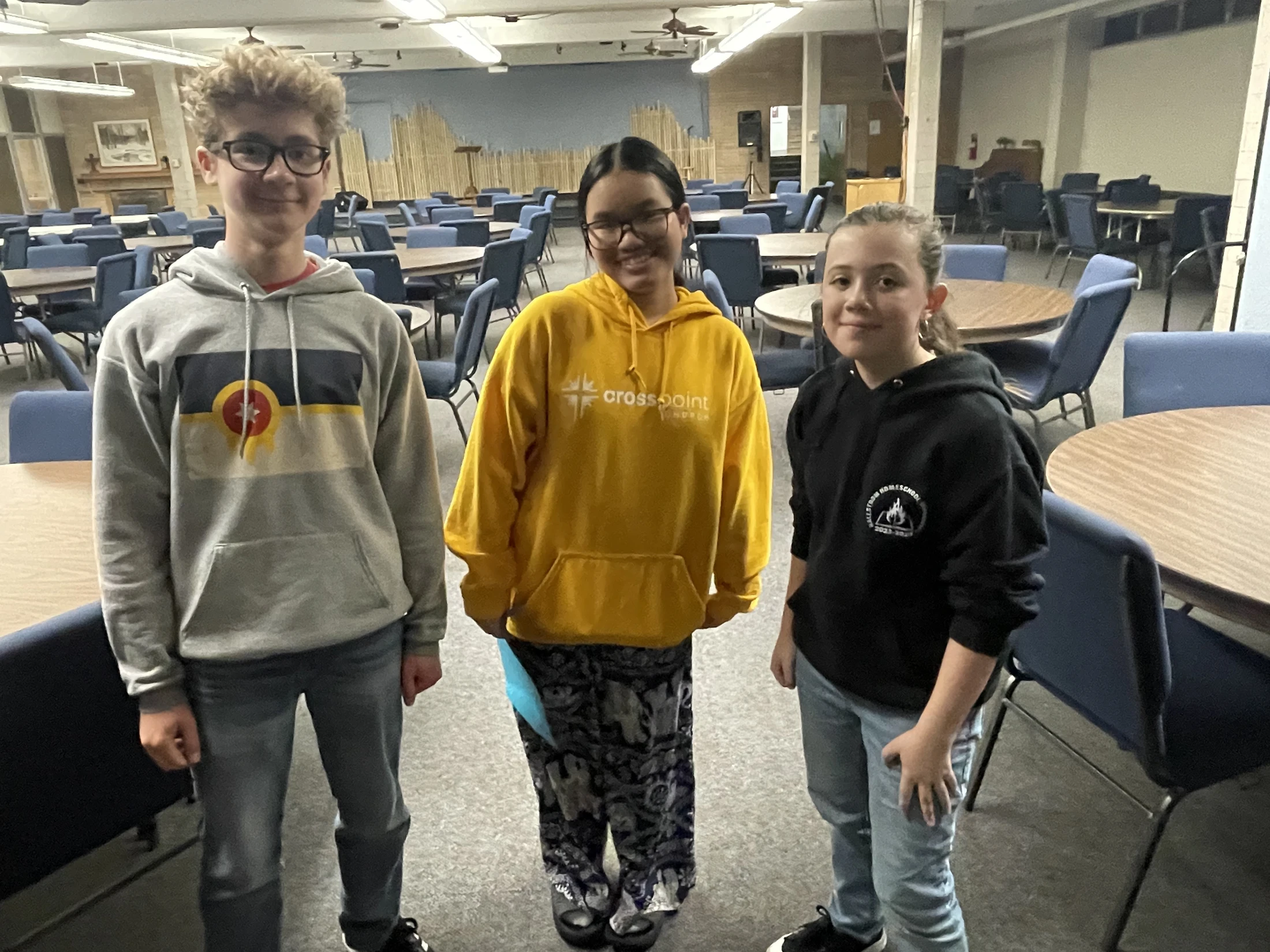 Three youths, a boy and two girls, stand smiling for a photo inside a large room filled with empty round tables and blue chairs.