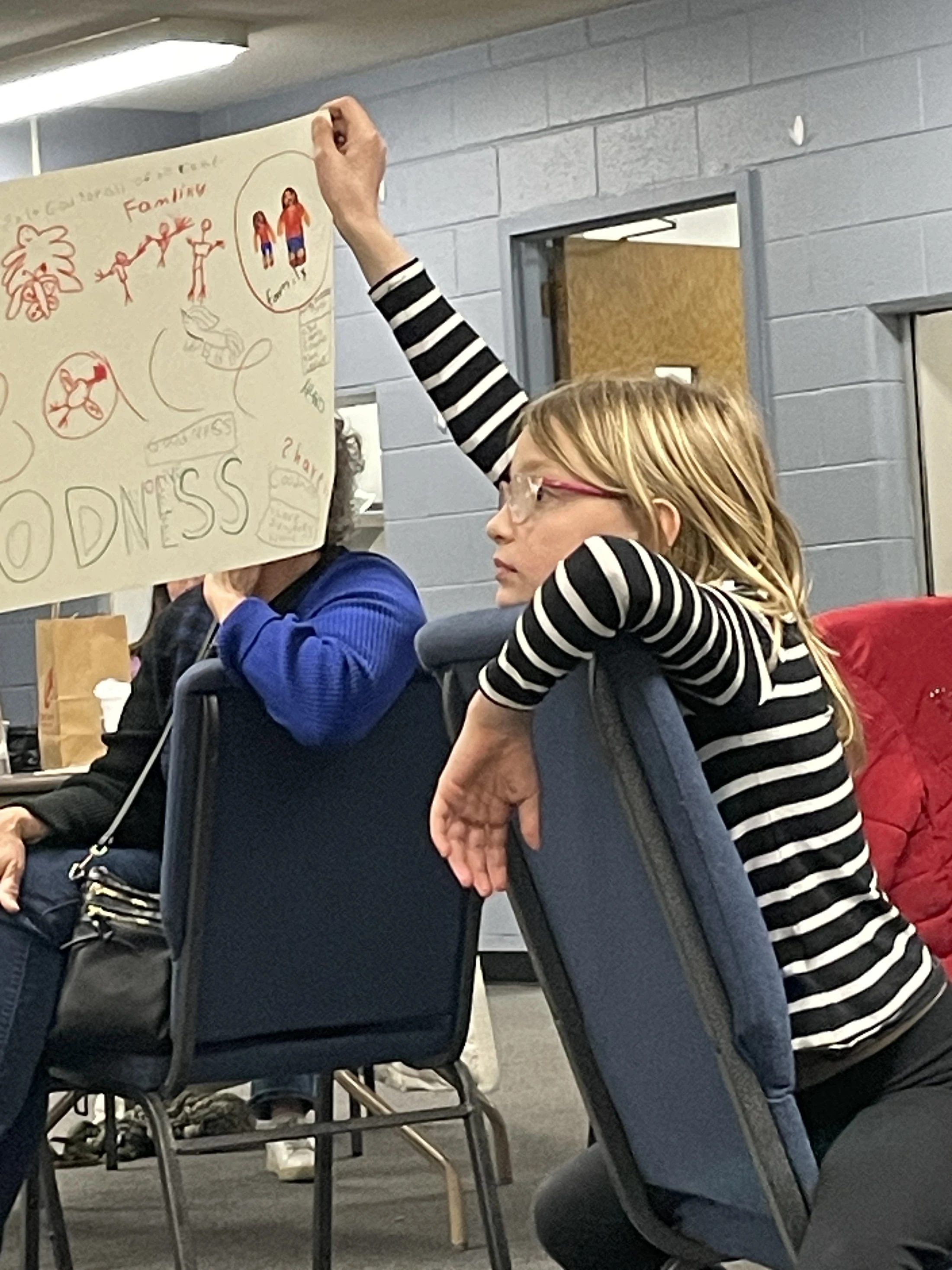 A young girl with glasses and a striped shirt sits in a chair, holding up a hand-drawn poster with the words 'Family' and 'GOODNESS' on it.