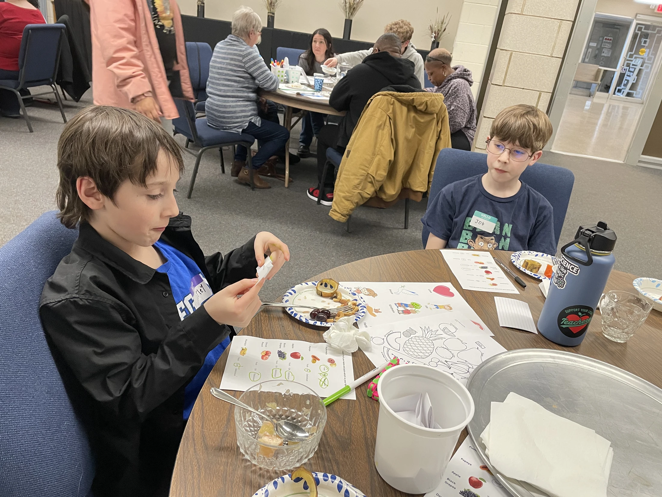 Two young boys sit at a round table with coloring sheets and snacks during a group event. One boy focuses on something in his hands while the other watches.