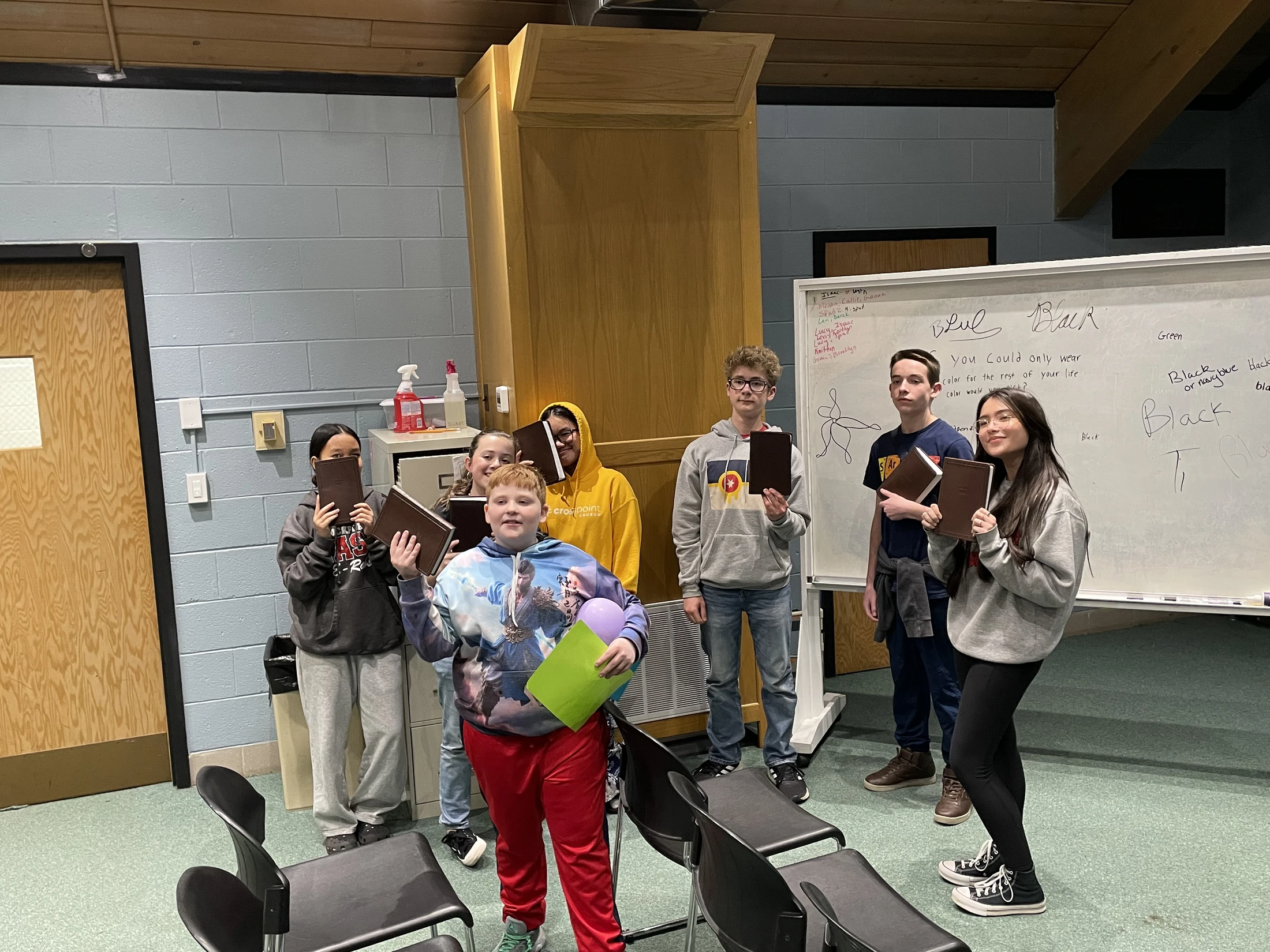 A group of seven young students stand together in a classroom, posing for a photo. Most are holding up identical brown, hardbound books.