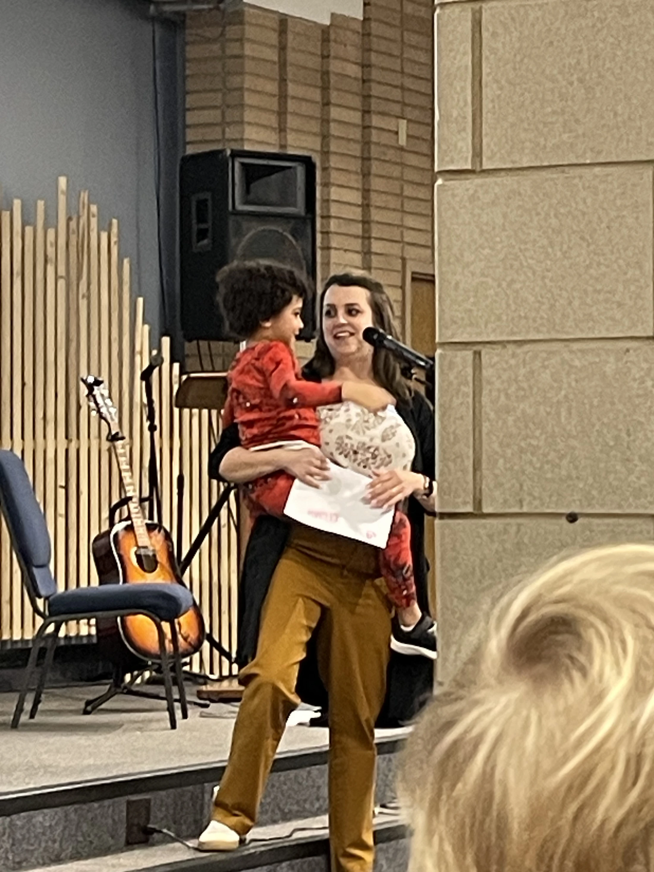A woman stands on a stage holding a small, smiling child in red pajamas. She is speaking into a microphone, and an acoustic guitar is in the background.