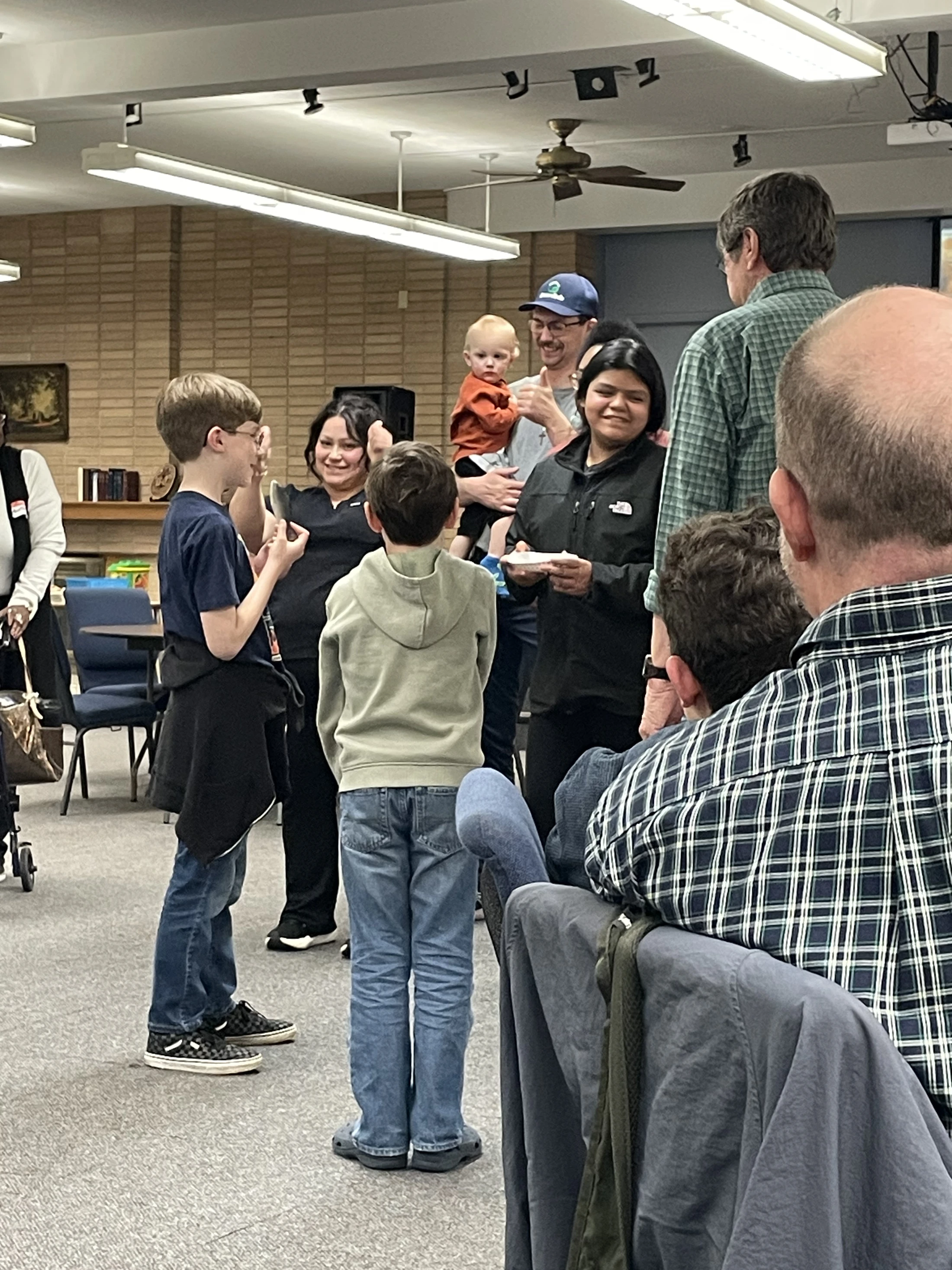 A diverse group of adults and children standing and talking in a well-lit community room. A man holds a baby, and a woman holds a paper plate.